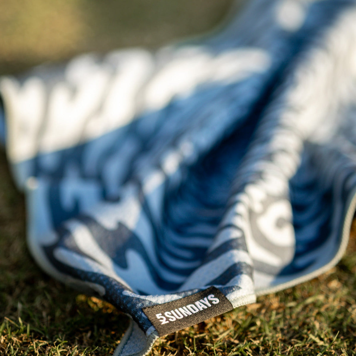 A close-up of a blue and grey patterned golf towel on grass, with a small black label reading “#SUNDAYS” visible near the edge.