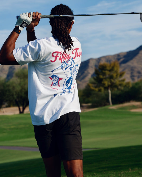 Golfer in the Be My Caddie Tee mid-swing, back graphic and "Fifty Two" script visible, desert course in the background.