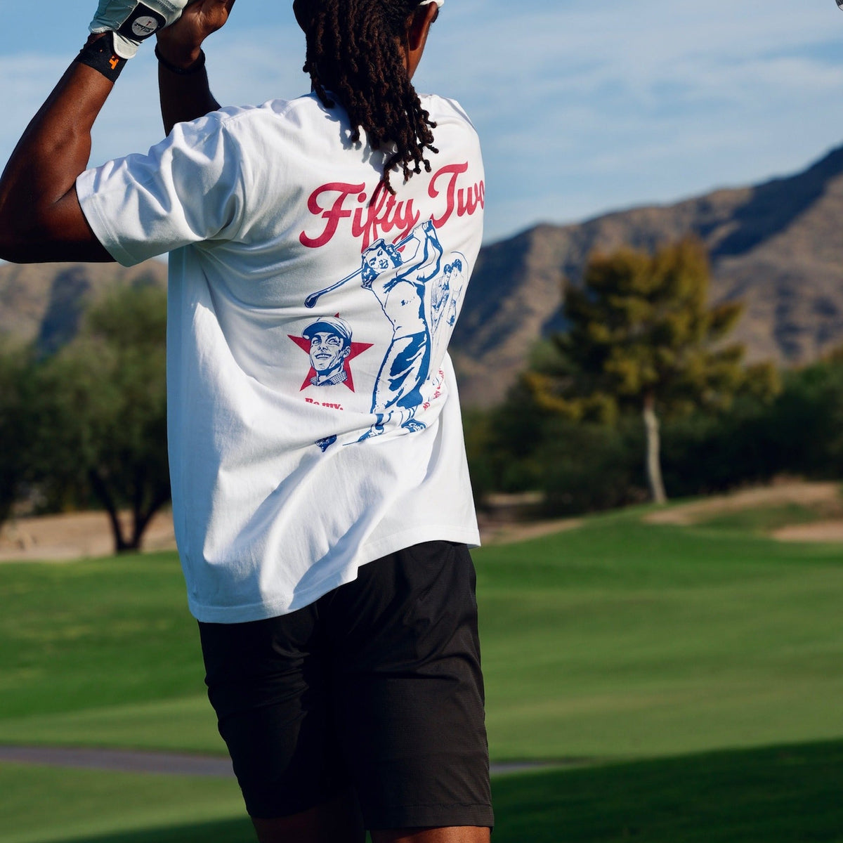 Golfer in the Be My Caddie Tee mid-swing, back graphic and "Fifty Two" script visible, desert course in the background.
