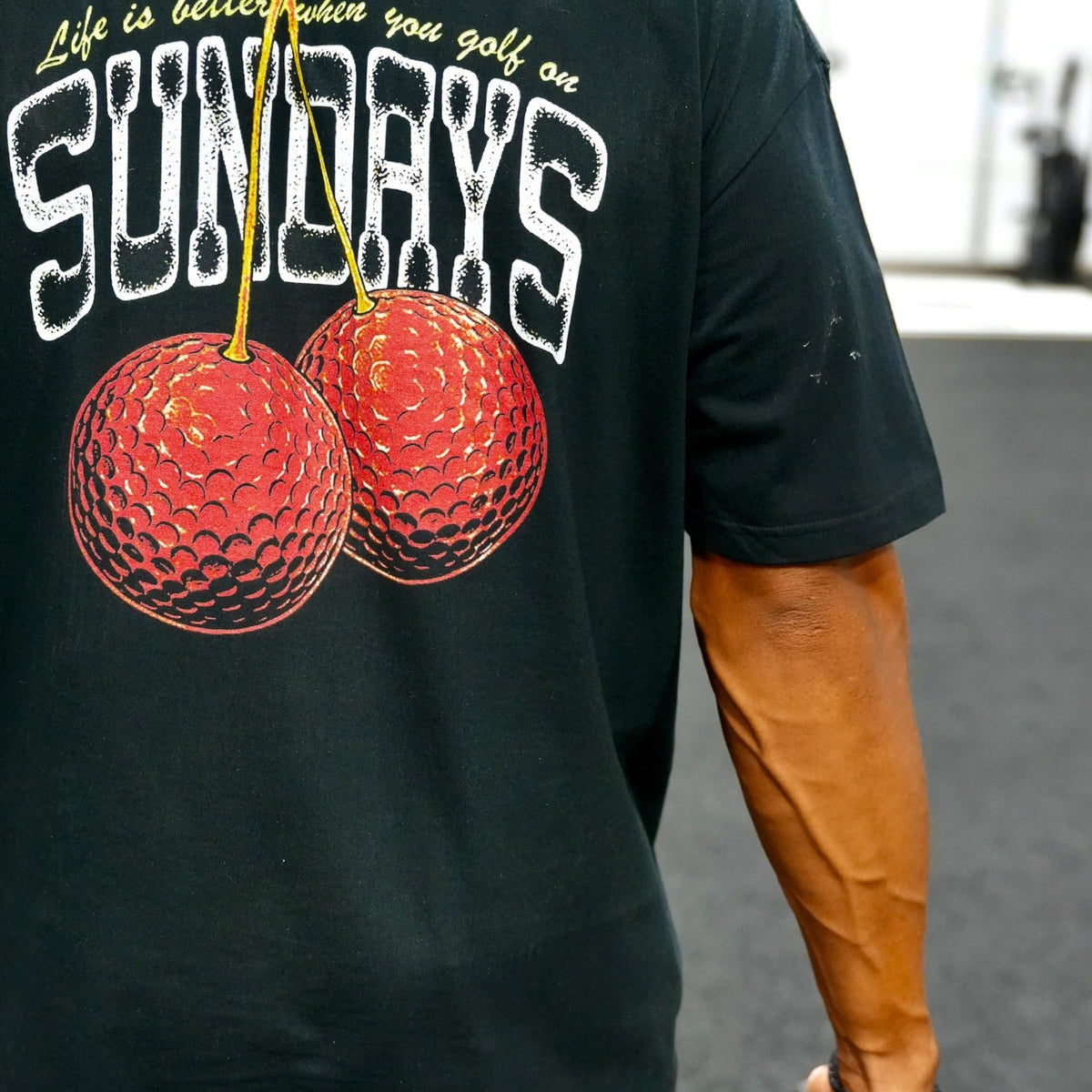 Close-up of the Sunday Cherries Tee's back graphic — two cherries made of golf balls with "Life is better when you golf on Sundays."