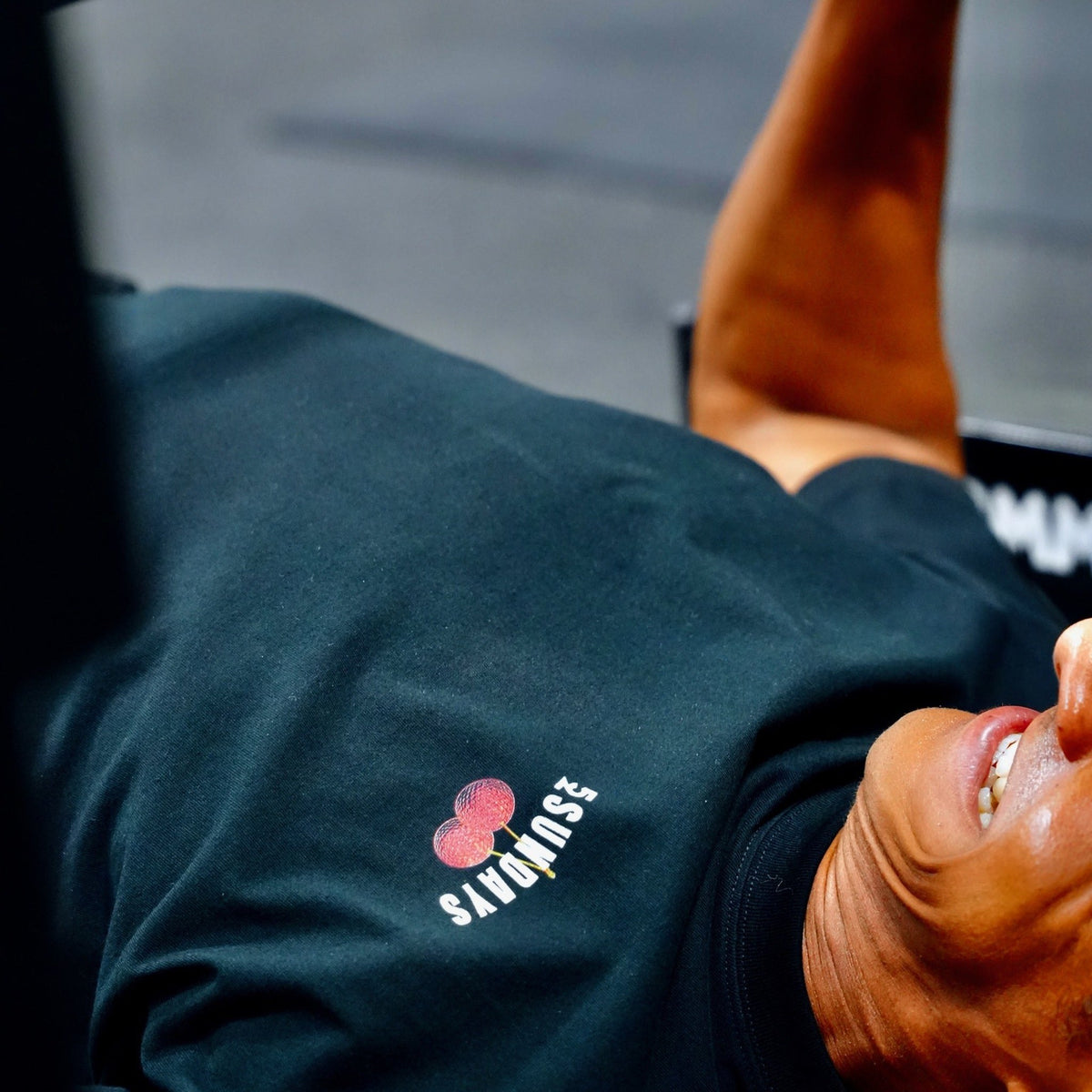 Close-up of the small front "52SUNDAYS" cherries logo on the Sunday Cherries Tee, worn during a bench press.