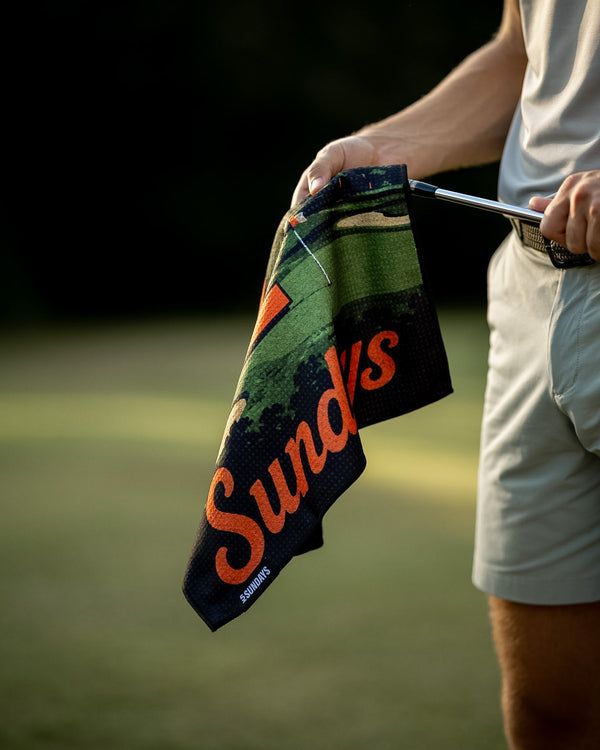 A close-up lifestyle shot of a person on a golf course holding a black towel with a golf course pattern and the orange text 'Sundays'
