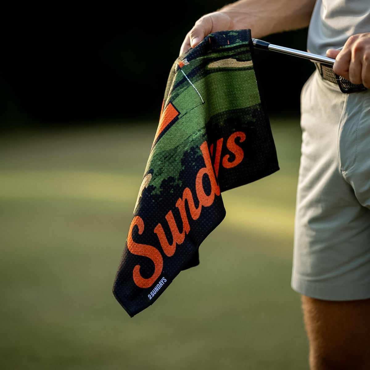 A close-up lifestyle shot of a person on a golf course holding a black towel with a golf course pattern and the orange text 'Sundays'