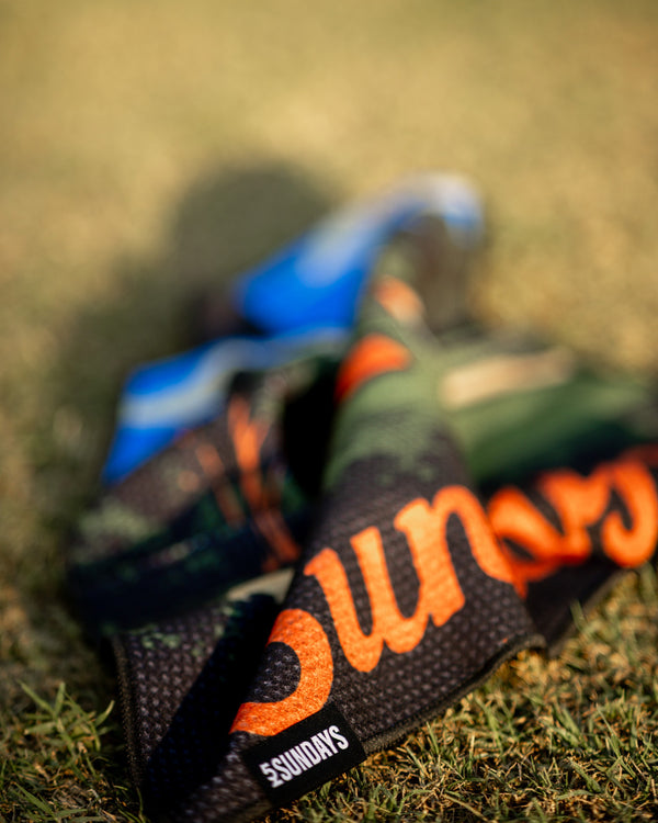 Close-up of 52 Sundays golf towels in a  golf course pattern print with orange script lettering, laid on grass.