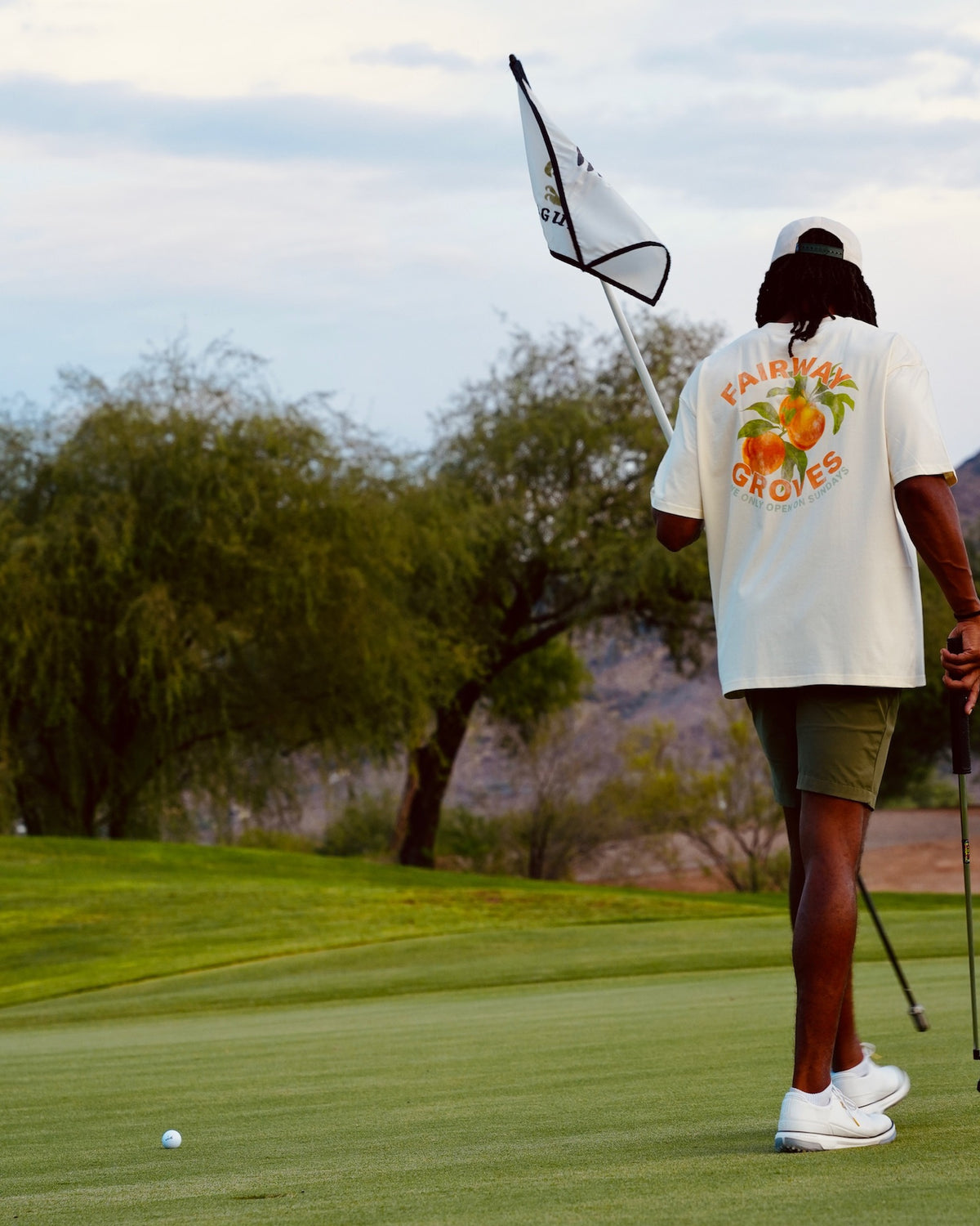Golfer wearing the Fairway Groves Tee on the green, holding the flagstick, back graphic visible.