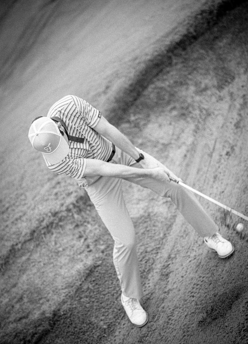 Golfer in a sand trap with a club, wearing a striped shirt and cap.