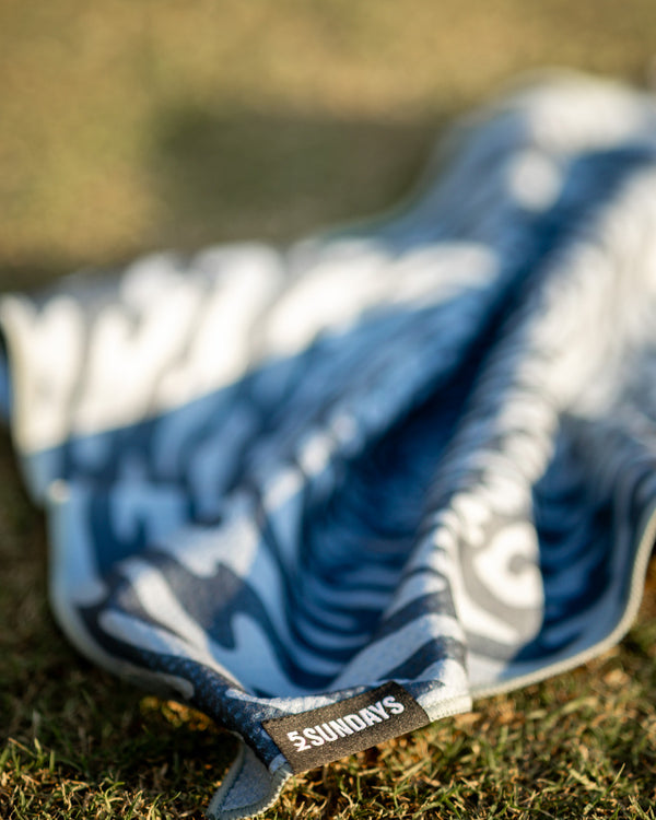 A close-up of a blue and grey patterned golf towel on grass, with a small black label reading “#SUNDAYS” visible near the edge.