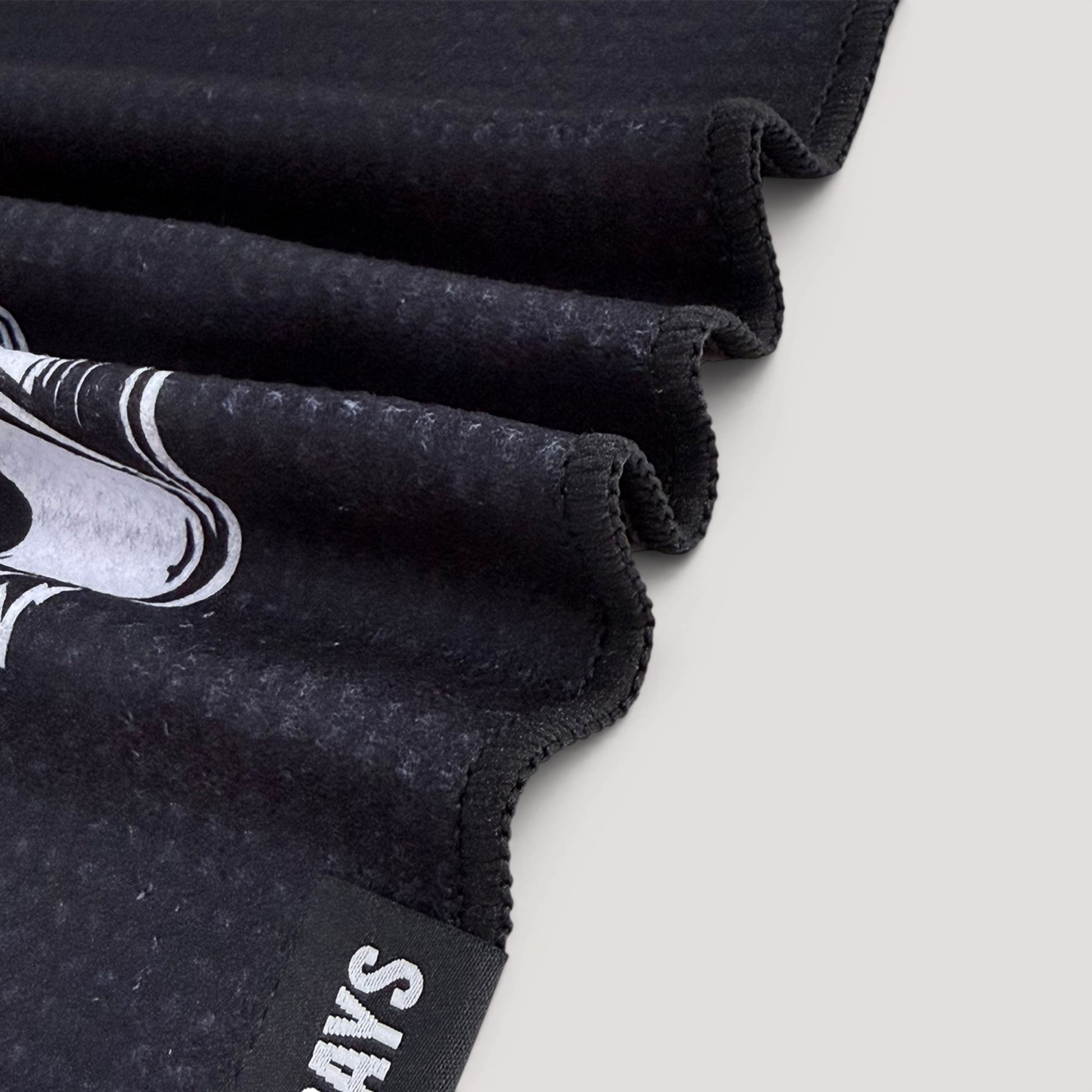 Close-up of a black textured fabric item, golf towel folded diagonally on a light gray background. The fabric features a white graphic print and a black fabric tag with white text.