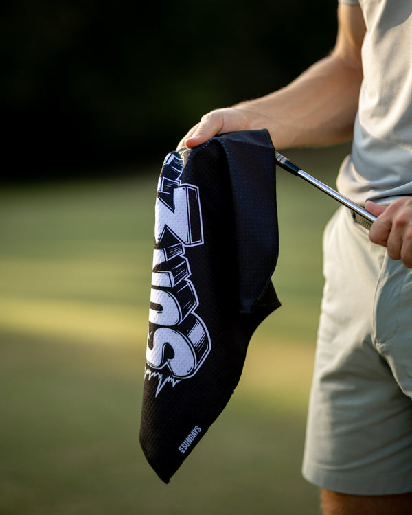 A close-up of a person outdoors holding a black golf towel with a large white and gray 'SUNDAYS' logo. 