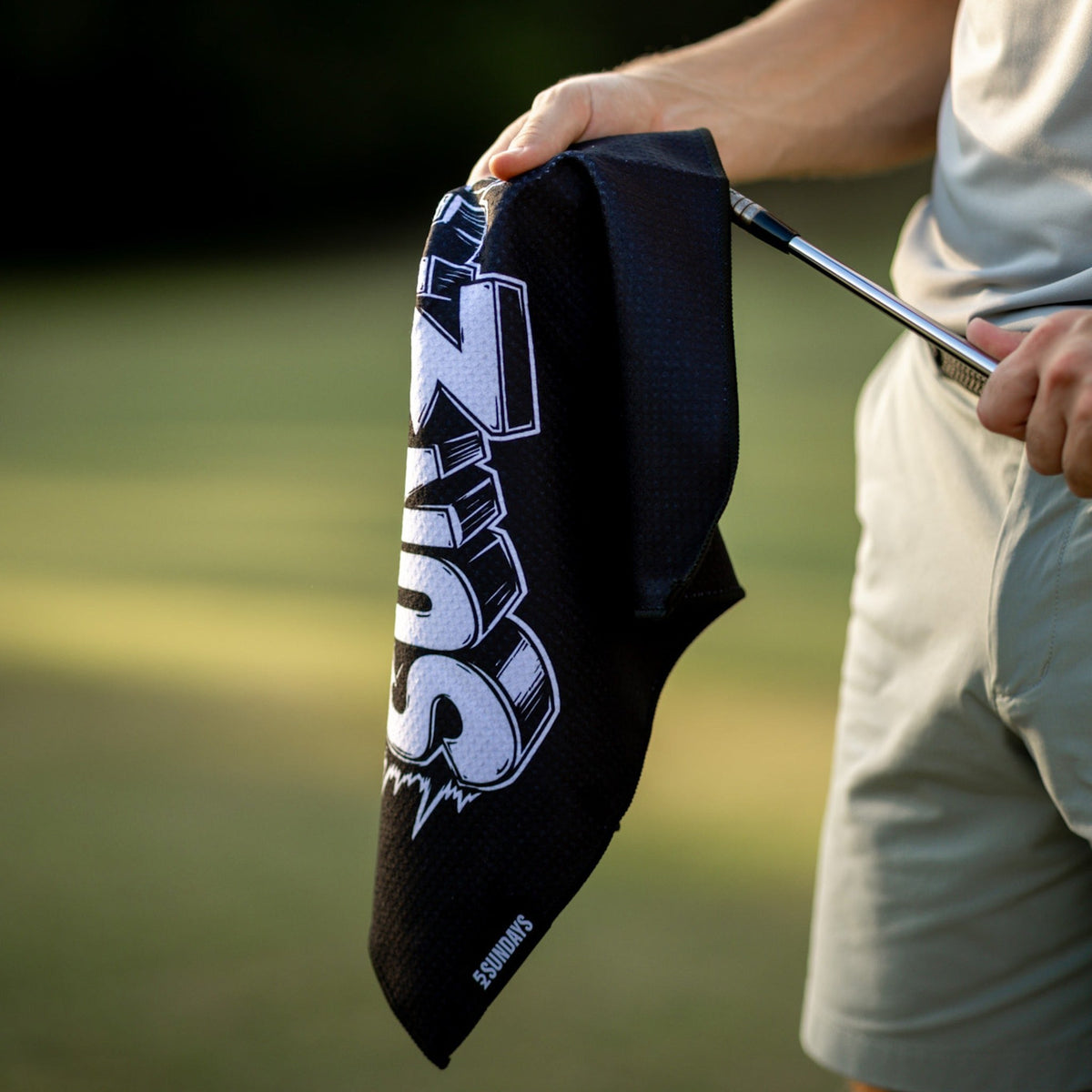 A close-up of a person outdoors holding a black golf towel with a large white and gray 'SUNDAYS' logo.