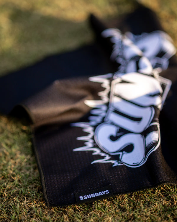 A black golf towel with a large white 'SUNDAY' graphic and '52 SUNDAYS' logo, laid out on green grass.