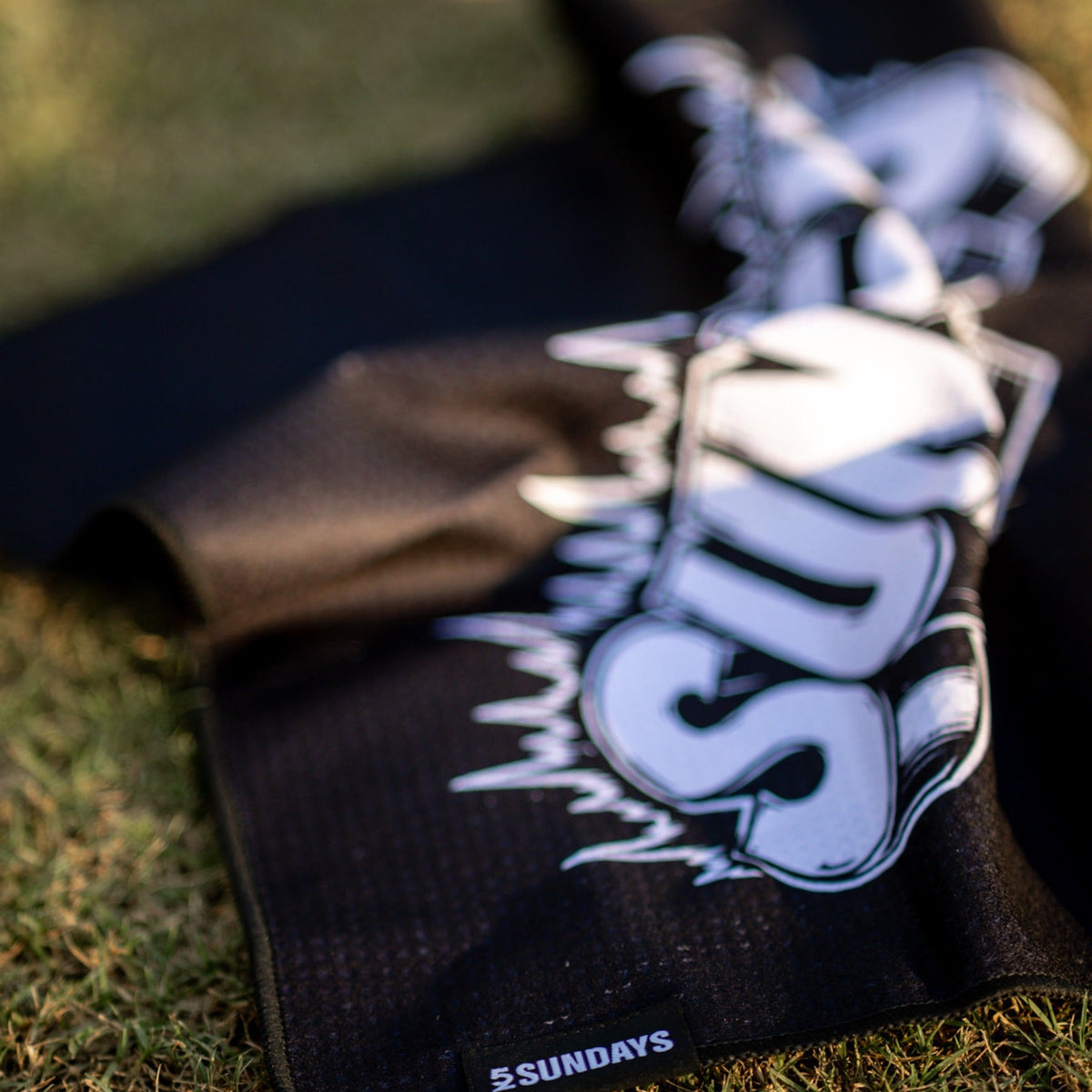 A black golf towel with a large white 'SUNDAY' graphic and '52 SUNDAYS' logo, laid out on green grass.